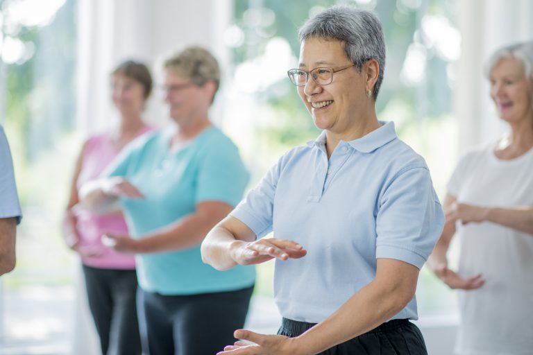 A multi-ethnic group of senior adults are taking a tai chi fitness class. They are practicing their martial art inside of a brightly lit studio. Here, a woman of asian ethnicity is leading the class through movements.
