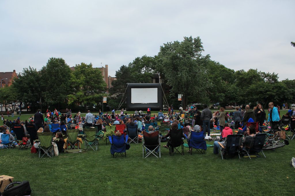 crowds sit in lawn chairs in front of a giant movie screen at scoville park