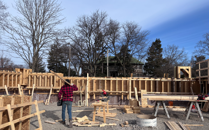 laborer in red shirt holds long beam of wood over shoulder while looking at wall frames
