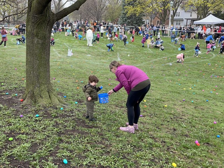 a mom hands her son an egg to add to his basket with dozens of kids running around in the background finding eggs