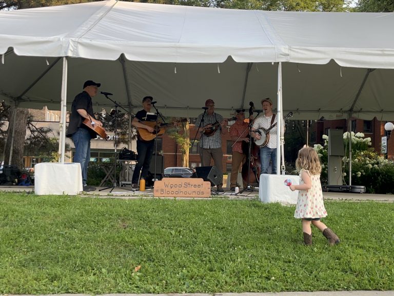 a girl in a white dress and cowgirl boots watches a band under a tent holding guitars