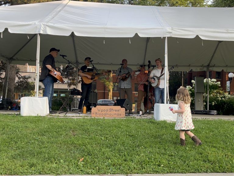 a girl in a white dress and cowgirl boots watches a band under a tent holding guitars