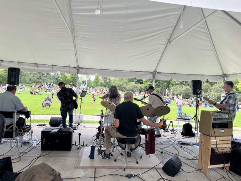 image of a band from behind with people holding guitars, sitting at a drum set, and singing