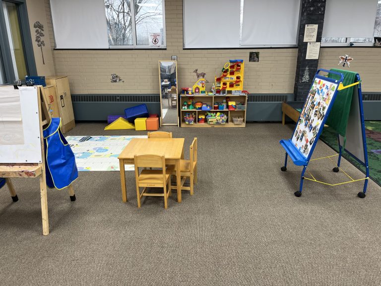 toddler sized table next to an easel covered in magnets and a toy shelf in the background